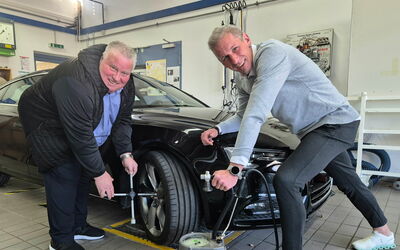Zwei Männer in der Werkstatt halten Radkreuz und Kompressor, posieren symbolisch für einen Reifenwechsel an einem Auto.