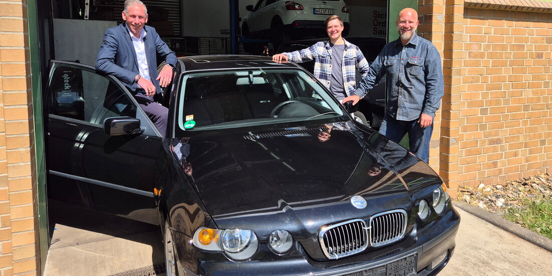 Drei Männer stehen links und rechts neben einem gespendeten Auto vor einer Werkstatt und präsentieren es für die Ausbildung.