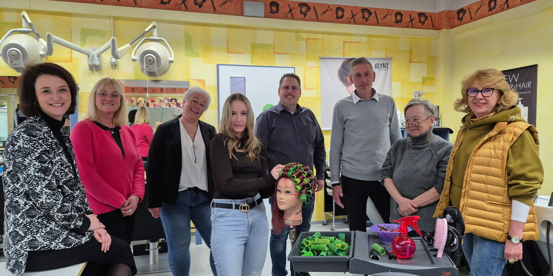 In der Friseurwerkstatt des BBZ in Korbach stehen die Netzwerkpartner des Projekts im Halbkreis um eine junge Fachpraktikerin im Friseurhandwerk im 1. Ausbildungsjahr. Die Fachpraktikerin dreht an einem Modellkopf Lockenwickler auf.